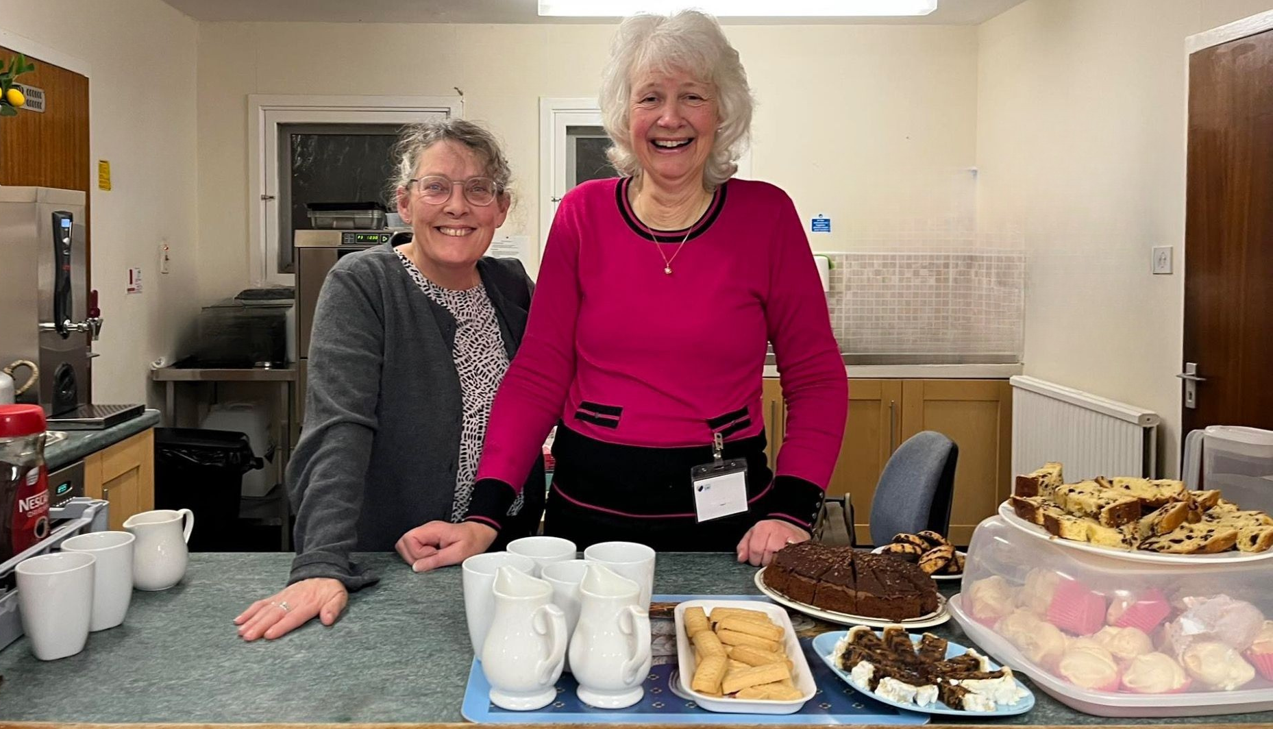 Two smiling Largs SWI members welcoming visitors with refreshments.